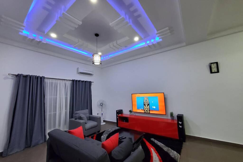 A modern living area features a stylish grey sofa adorned with red cushions. A large television is mounted on a red media console, complemented by dark curtains and a lighted ceiling. A fan adds comfort in the space, with artwork and a clock enhancing the decor.