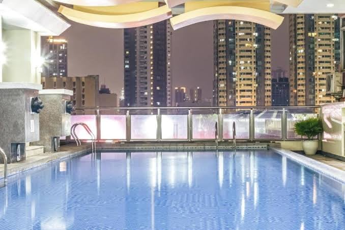 The rooftop pool is surrounded by a glass railing, providing views of the city skyline at night. Ambient lighting reflects off the water, creating a serene atmosphere. The pool area features a smooth surface with a staircase for easy access.