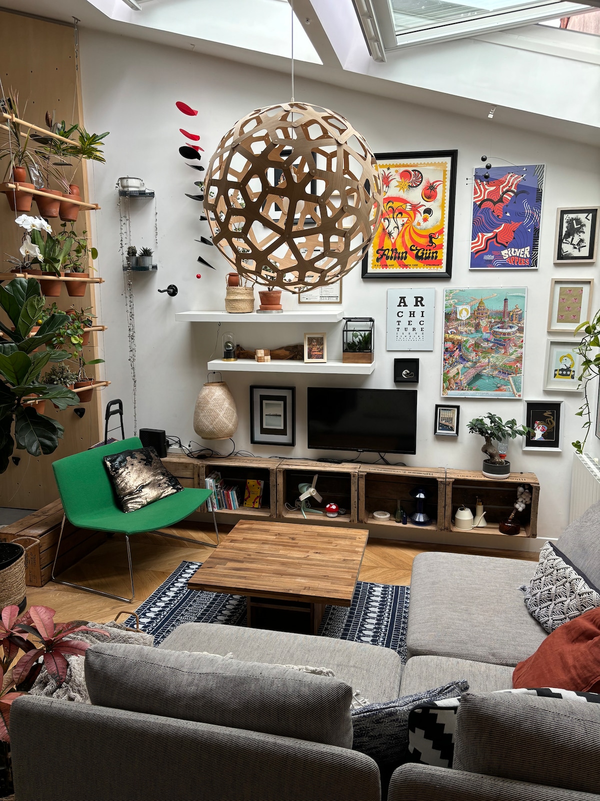 The living area features a bright atmosphere with a skylight above. A wooden coffee table sits at the center, surrounded by plush seating in neutral tones. Vibrant artwork and decorative shelves adorn the walls, complemented by greenery throughout the space and a unique light fixture above.