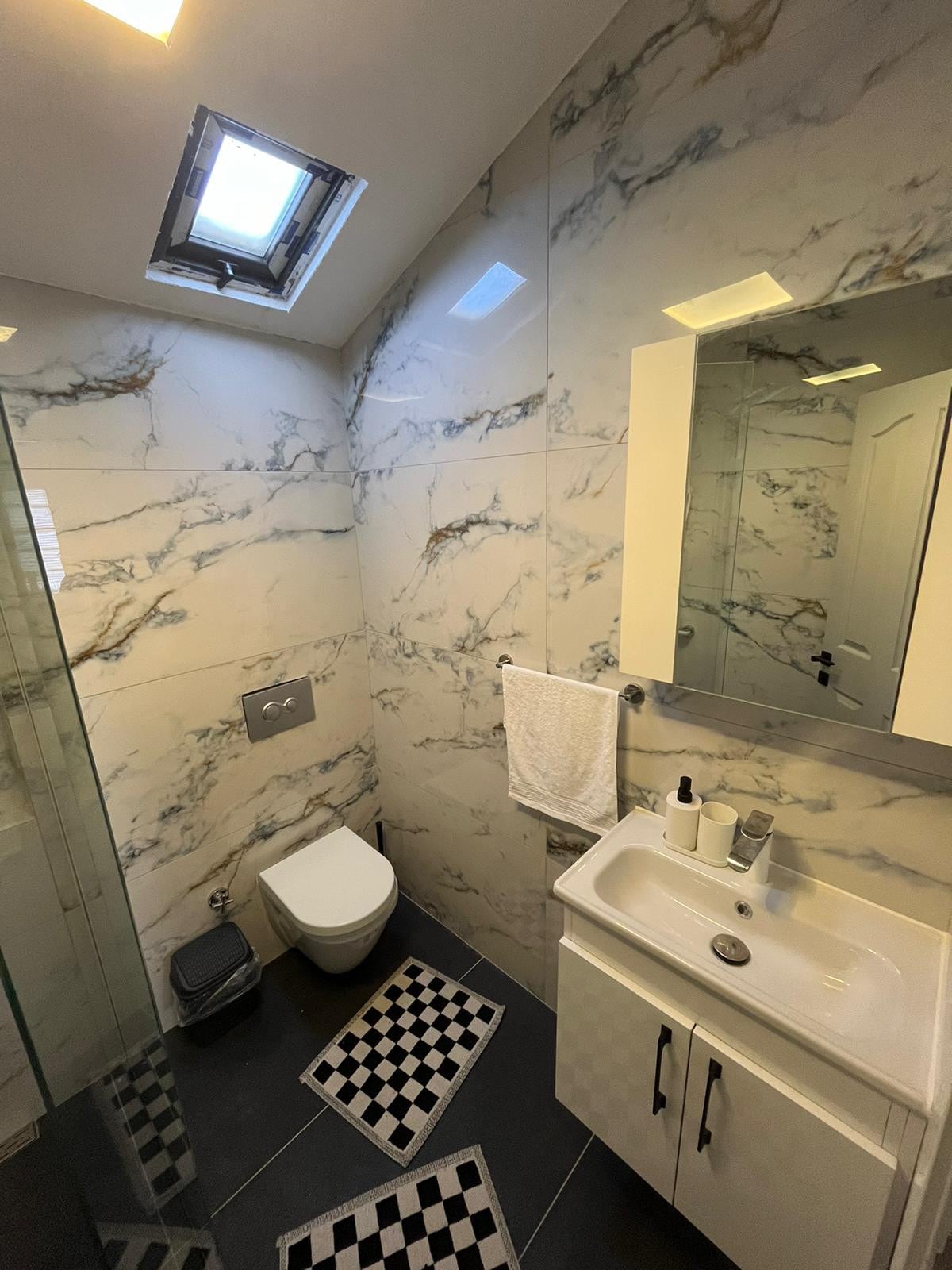 A modern bathroom features elegant marble walls and a skylight providing natural light. A sleek sink with a large mirror is positioned above storage cabinets. A stylish shower area is enclosed by glass, and a checkered mat adds a contemporary touch to the floor.