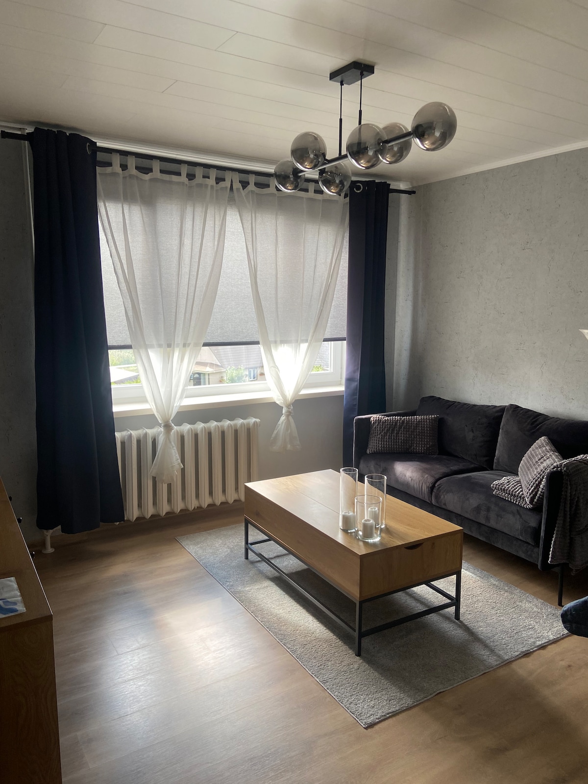 A modern living area is presented, featuring a dark sofa beside a wooden coffee table with decorative glass vases. Natural light is filtered through sheer window curtains, and a sleek light fixture hangs above, complementing the neutral color palette of the room.