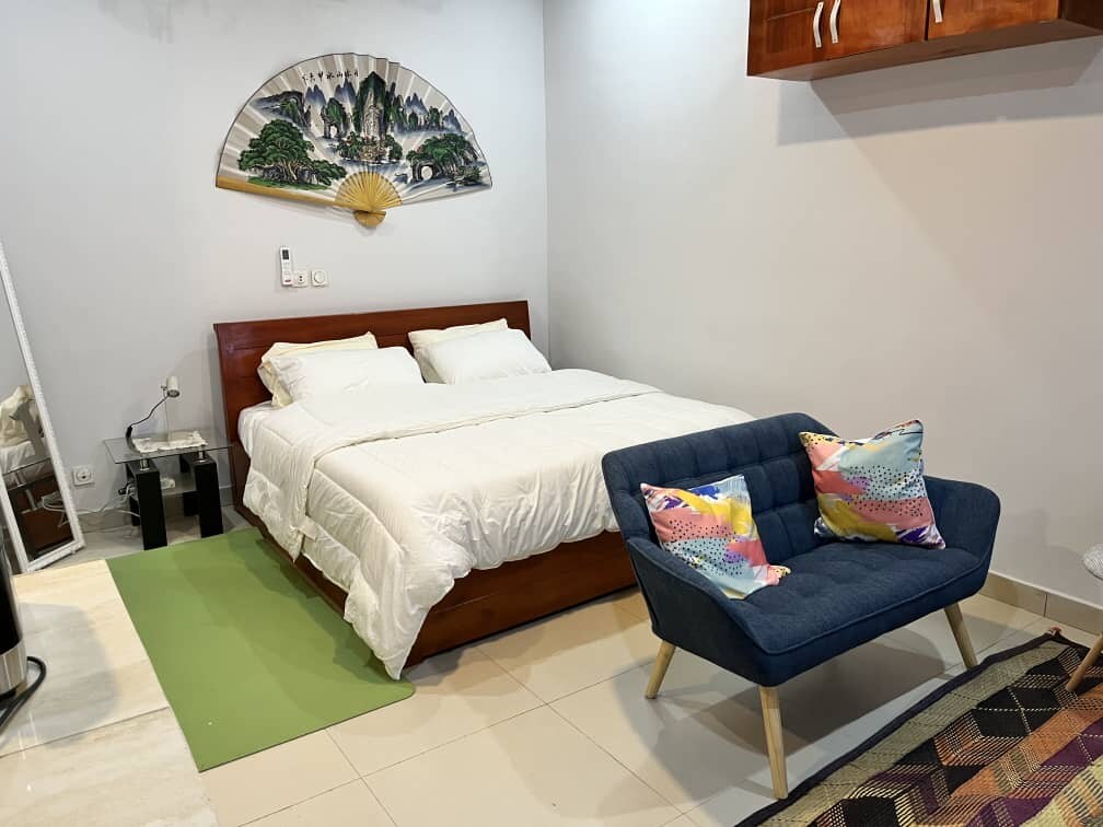 A well-appointed sleeping area features a spacious bed with white linens, complemented by a colorful accent chair nearby. A minimalist decorative fan adorns the wall, while natural light is enhanced by soft tones from the surrounding walls.