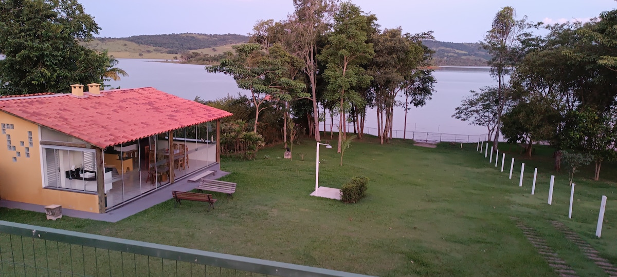 A spacious yard is set beside a serene lake, with well-maintained grass and scattered trees providing shade. A charming house with a red-tiled roof and large windows appears next to the water, offering outdoor seating areas for relaxation.