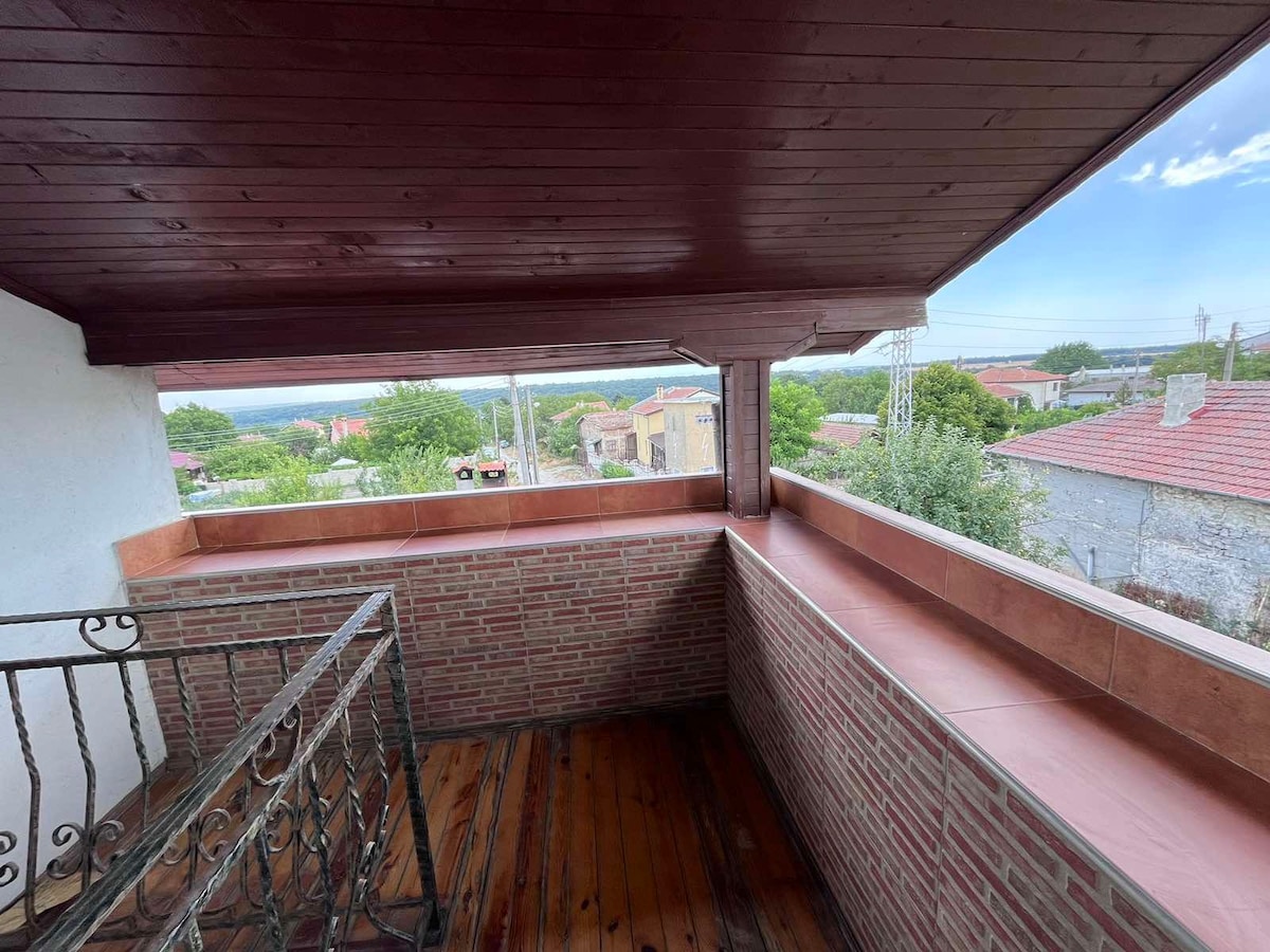 A private balcony features wooden flooring and brick walls, offering a spacious area for relaxation. The balcony overlooks the surrounding landscape, which includes greenery and nearby buildings. A wooden railing adds a rustic touch to the space, complemented by a wooden ceiling overhead.