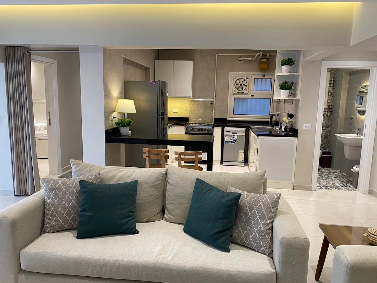 An open living space features a light-colored sofa adorned with two dark green cushions. The modern kitchen area is visible, showcasing stainless steel appliances and a sleek countertop. Natural light illuminates the room, highlighting the neutral tones and minimalist design.