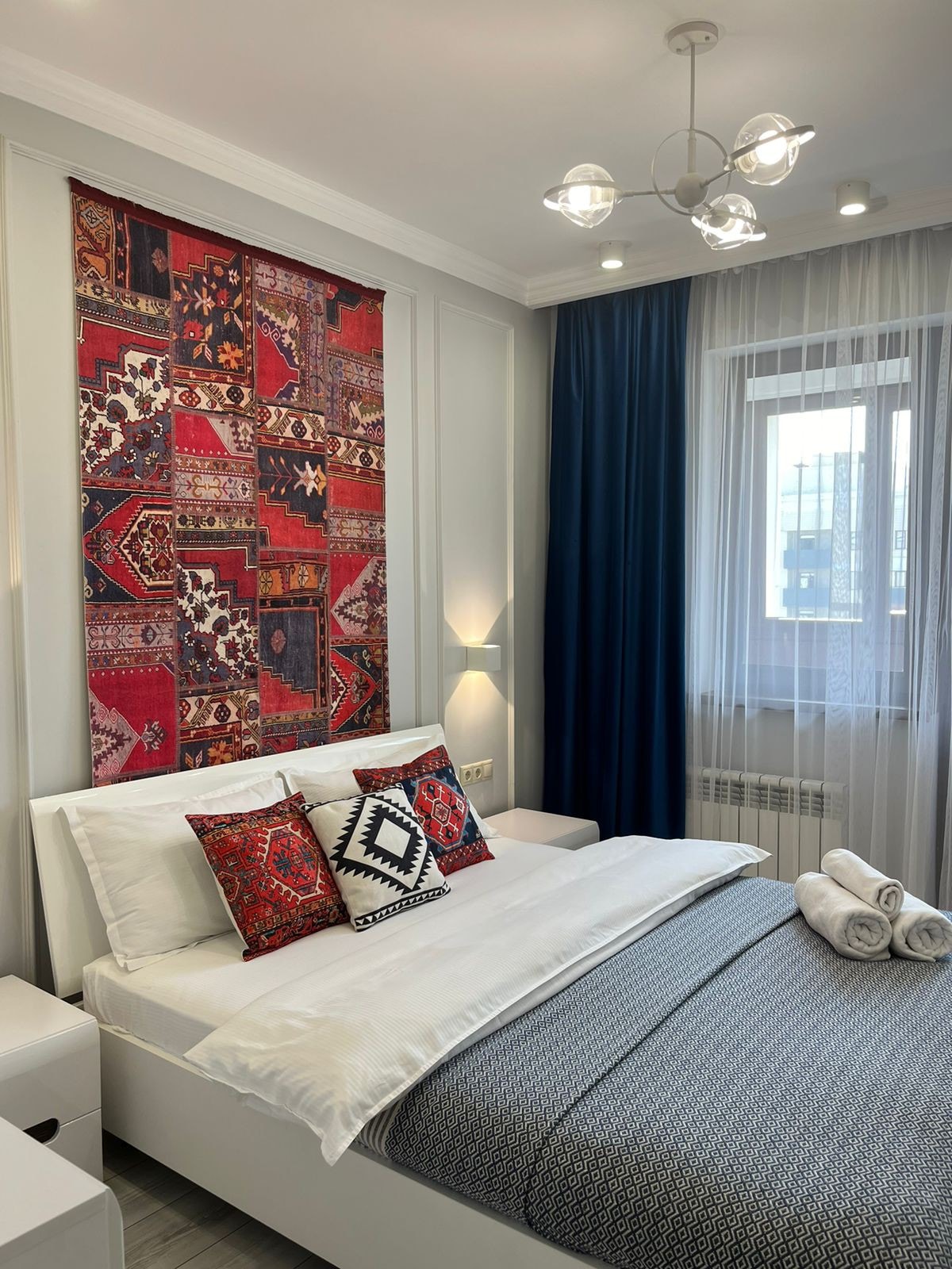 A bright bedroom features a spacious bed adorned with decorative pillows and a soft blanket. A patterned tapestry hangs above the headboard, and natural light filters through sheer curtains. The room is complemented by modern lighting and a clean, minimalist aesthetic.