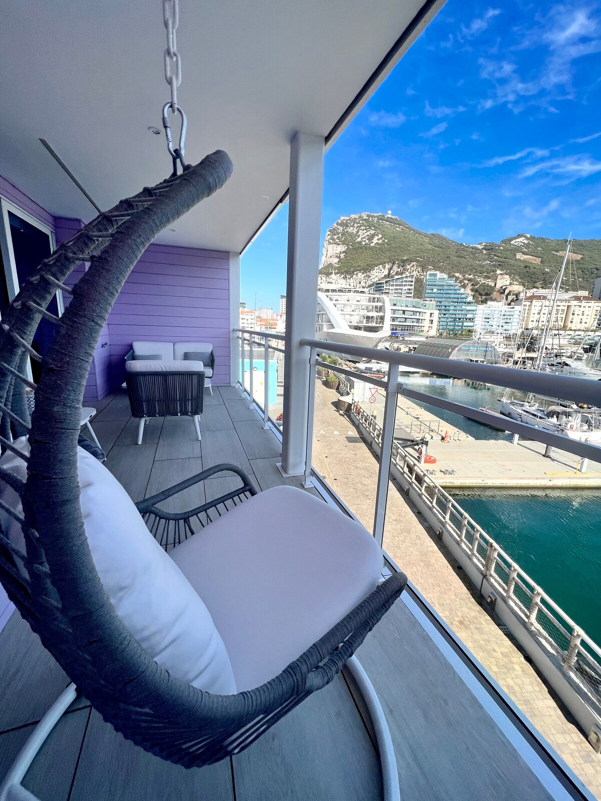 A hanging chair is positioned on a balcony, providing a view of the marina and surrounding buildings. The space features contemporary furnishings and a wooden deck, with gentle waves visible in the water below and the Rock in the background.