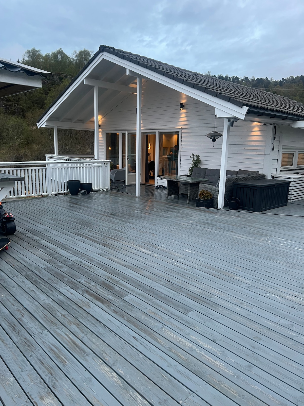 A spacious wooden deck is shown, featuring outdoor seating and a hot tub. The white house is partially visible, with large glass doors leading indoors. Soft lighting enhances the setting, complemented by surrounding greenery in the background.