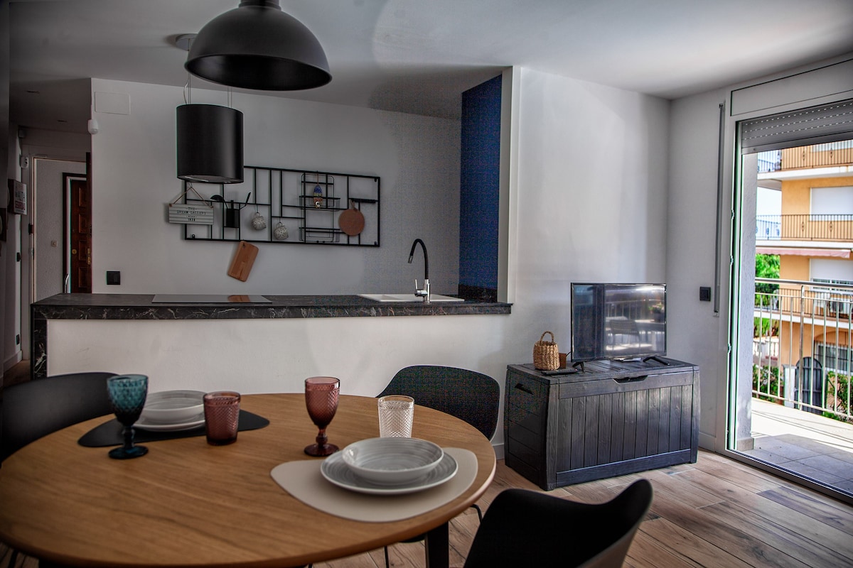 A modern dining area features a round wooden table set with dishes, glasses, and a warm ambiance. The kitchen area showcases a dark countertop and minimalist decor, while a sliding glass door provides access to a balcony, allowing natural light to fill the space.