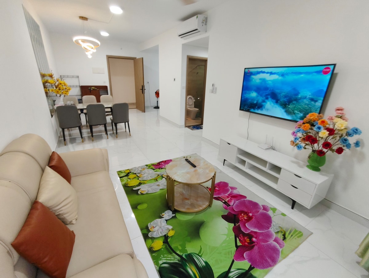A light-filled living area features a beige sofa flanked by decorative pillows. A floral rug complements a round coffee table, which holds a remote. A wall-mounted TV displays an underwater scene. In the background, a dining area with six chairs is visible.