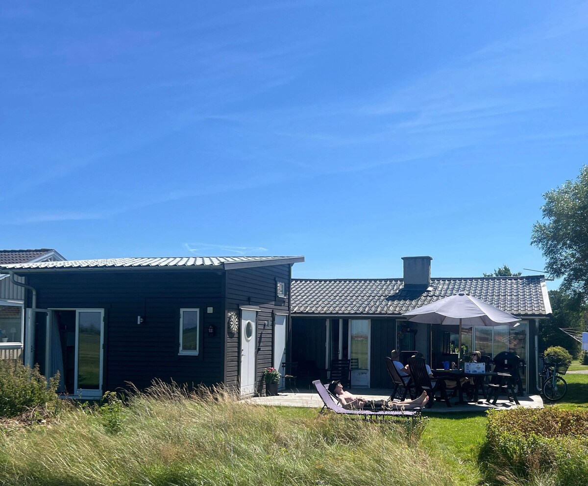 A modern single-story home features a dark exterior with large glass doors and windows, allowing natural light to fill the interior. A covered outdoor dining area is furnished with a table and several chairs, providing a space for family gatherings. Lush greenery surrounds the property.