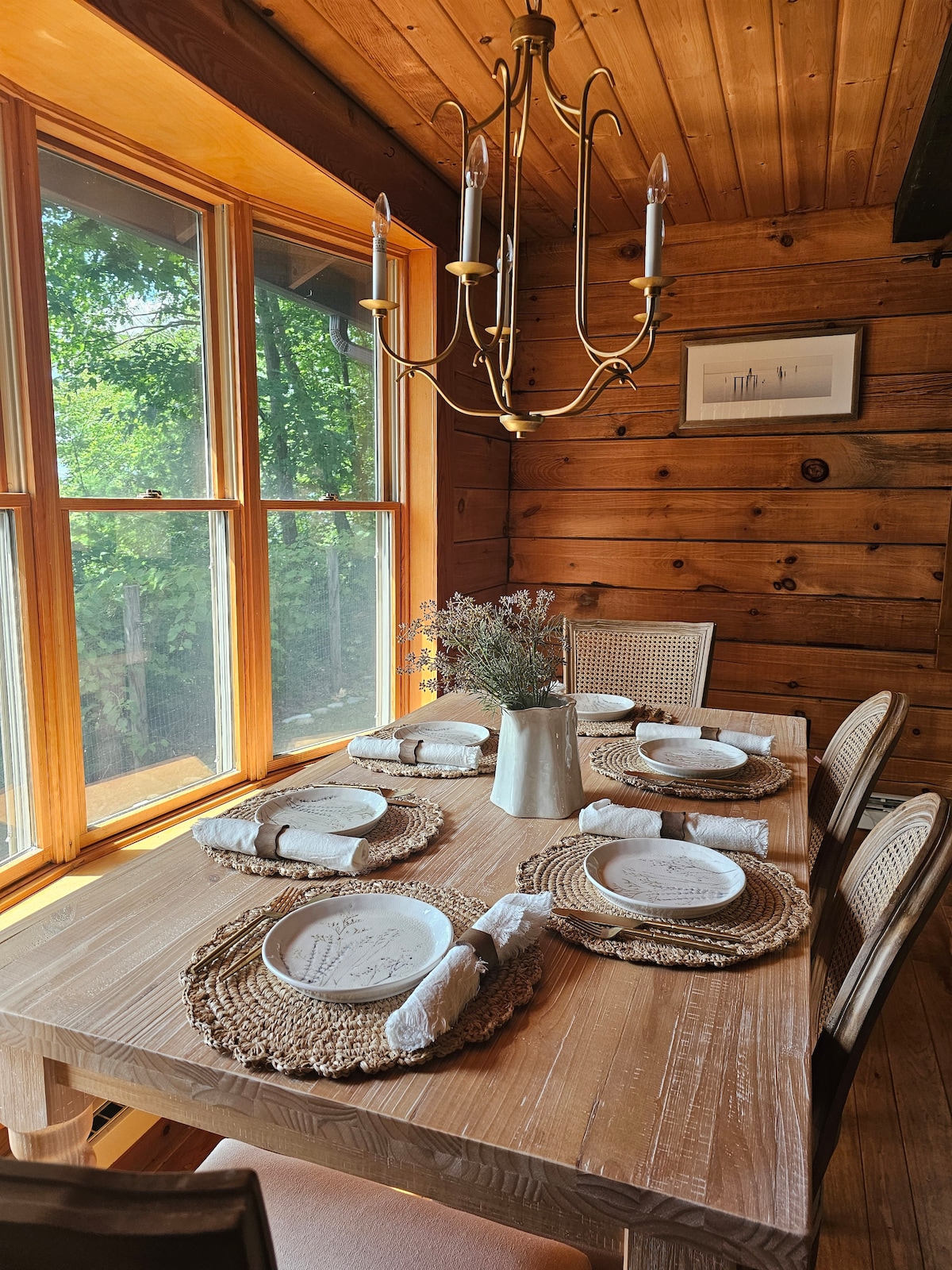 A wooden dining table is set for six, featuring woven placemats and white plates. Surrounding the table are light-colored chairs, and natural light pours in through large windows framed by wooden walls. A simple chandelier hangs above, adding a warm touch.