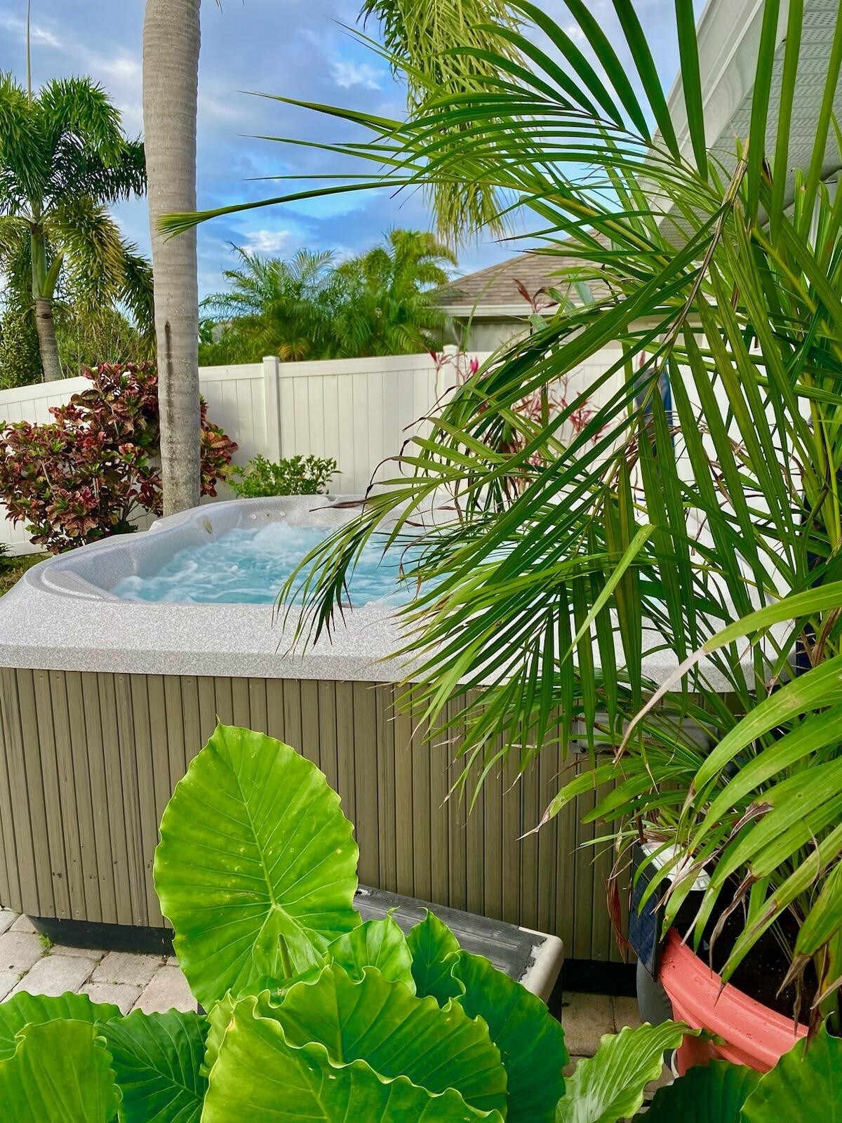 A hot tub is tucked into a private area, surrounded by lush greenery and potted plants. Palm trees and vibrant foliage create a tropical ambiance, while the water in the tub is gently rippling, reflecting the inviting surroundings.