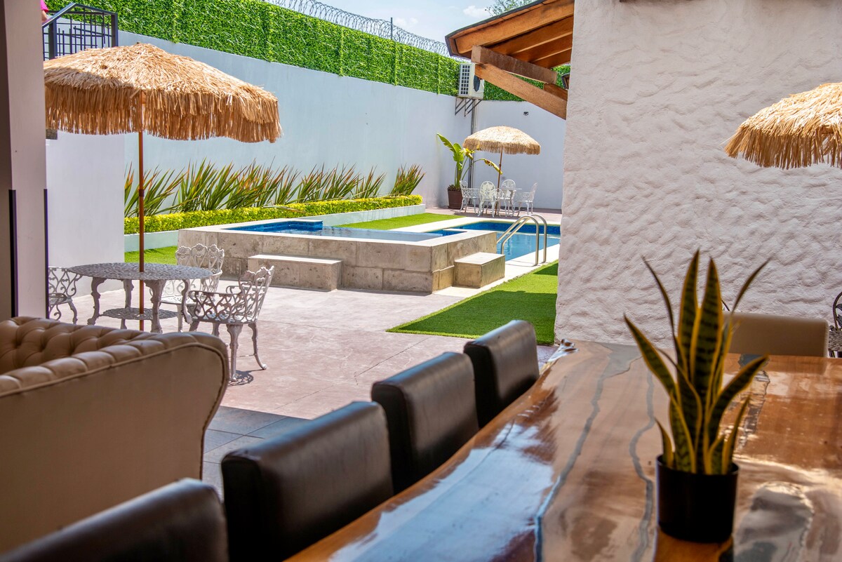 An inviting outdoor space features a swimming pool flanked by greenery and lounge areas. Straw umbrellas provide shade over seating arrangements, while a dining table is positioned indoors, showcasing a smooth wooden surface and comfortable black chairs. Light fills the area, enhancing the tranquil ambiance.