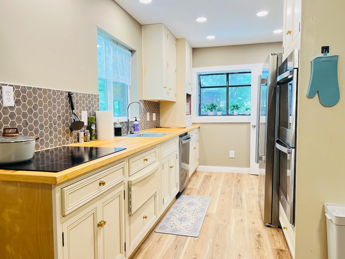 The kitchen features a modern design with light cabinetry and a wood countertop. Stainless steel appliances, including a refrigerator and dishwasher, are visible. A large window provides natural light and a view of the outdoors. The space is equipped with a stove and ample storage.