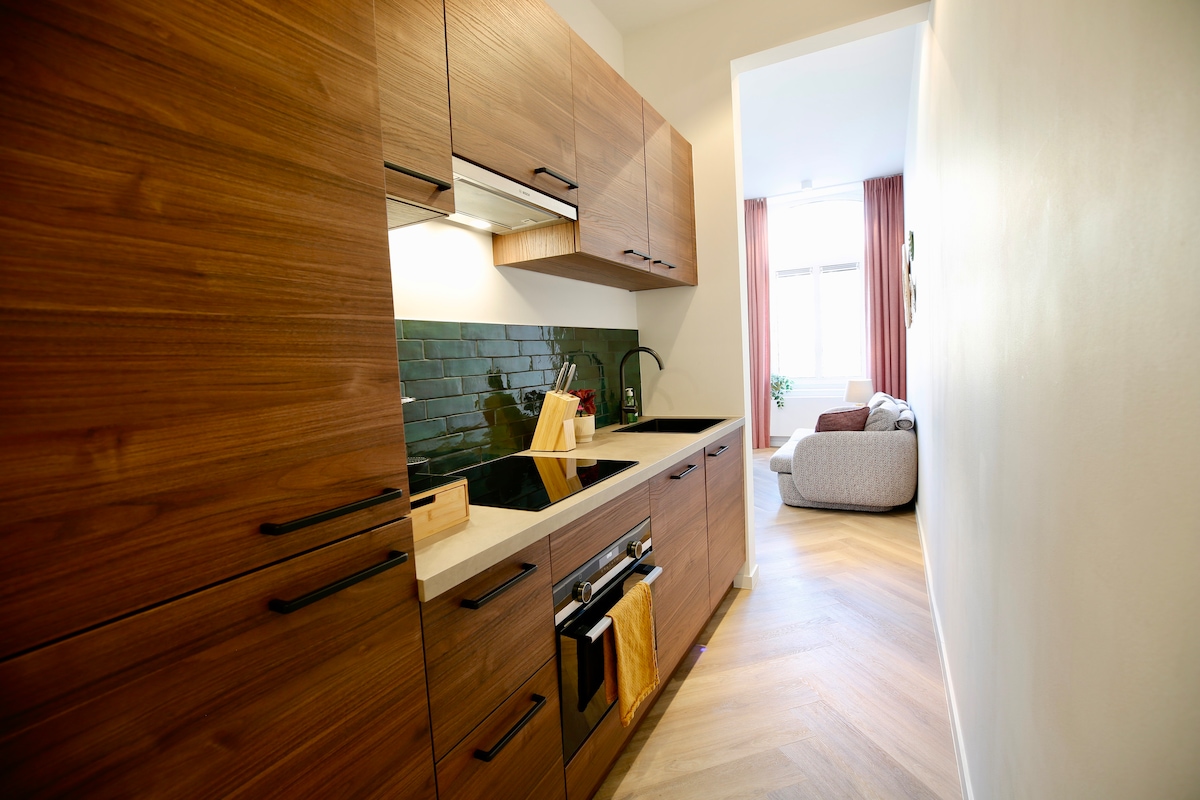 A modern kitchen features wooden cabinetry and sleek countertops. Appliances, including a stove and sink, are integrated into the design. A potted plant is visible through a window, while light filters in, creating a warm ambiance in the adjacent living area.