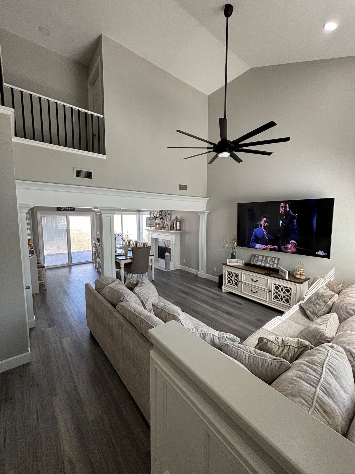 An open-concept living area features a large sectional sofa arranged around a coffee table. A smart TV is mounted on the wall, facing the seating. A ceiling fan hangs above, while a dining area with a table is visible through an archway in the background.