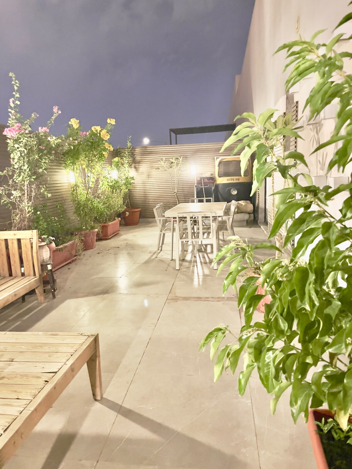 An outdoor patio is presented with wooden seating and a dining table surrounded by colorful potted plants. A gentle glow from ambient lighting creates a relaxing atmosphere, enhancing the tranquil appeal of the space.