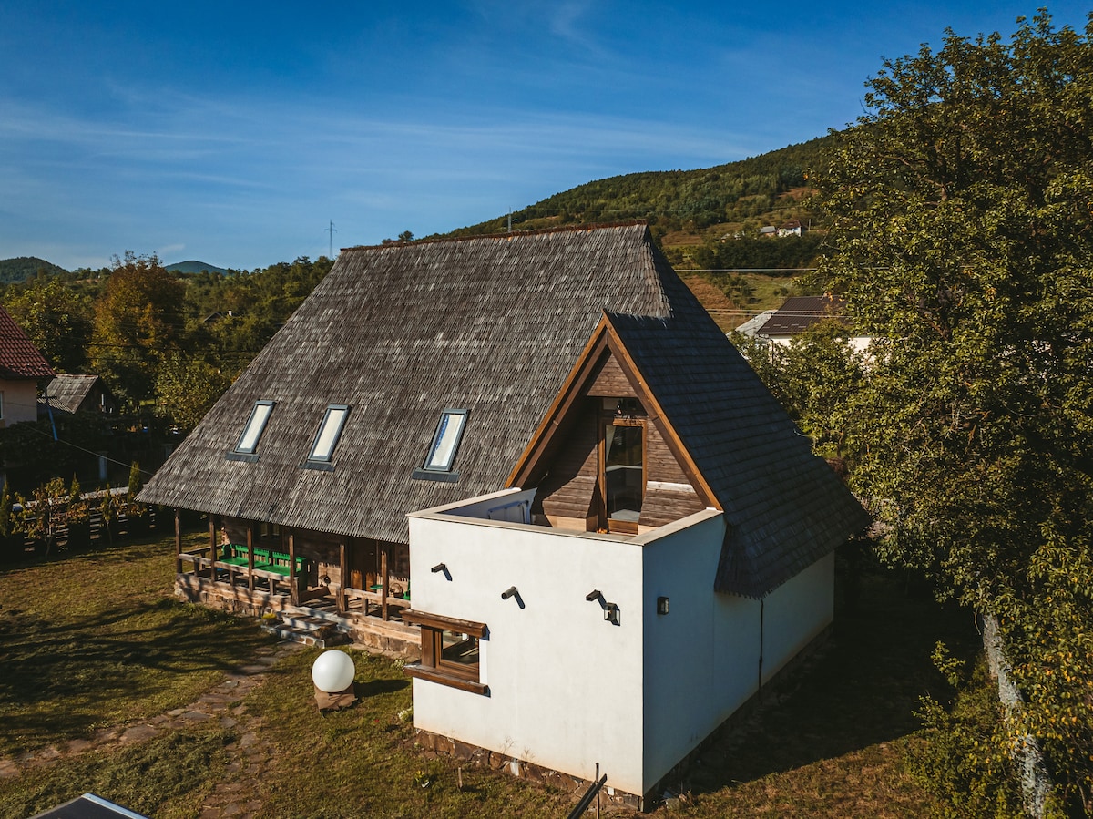 The traditional house features a thatched roof and a blend of wood and white stone exterior. Large windows are visible, allowing light to enter the spacious interior. Green hills and a clear blue sky create a serene backdrop.