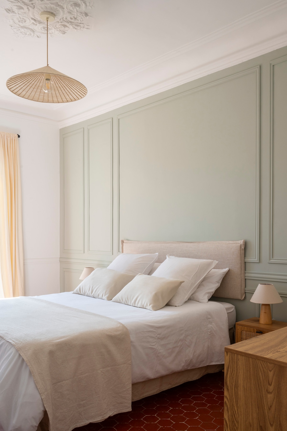 The bedroom features a spacious bed adorned with multiple beige pillows, set against a soft green accent wall. Two bedside tables, each with a lamp, complement the light-colored furnishings. Natural light filters through sheer curtains, enhancing the room's calming ambiance.