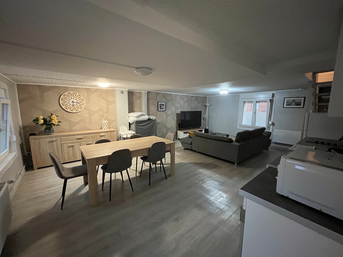The spacious living area features a modern dining table surrounded by four chairs, a comfortable sofa, and a television. Soft lighting enhances the welcoming atmosphere. Neutral tones are used on the walls, complemented by decorative elements, creating a functional and inviting space.