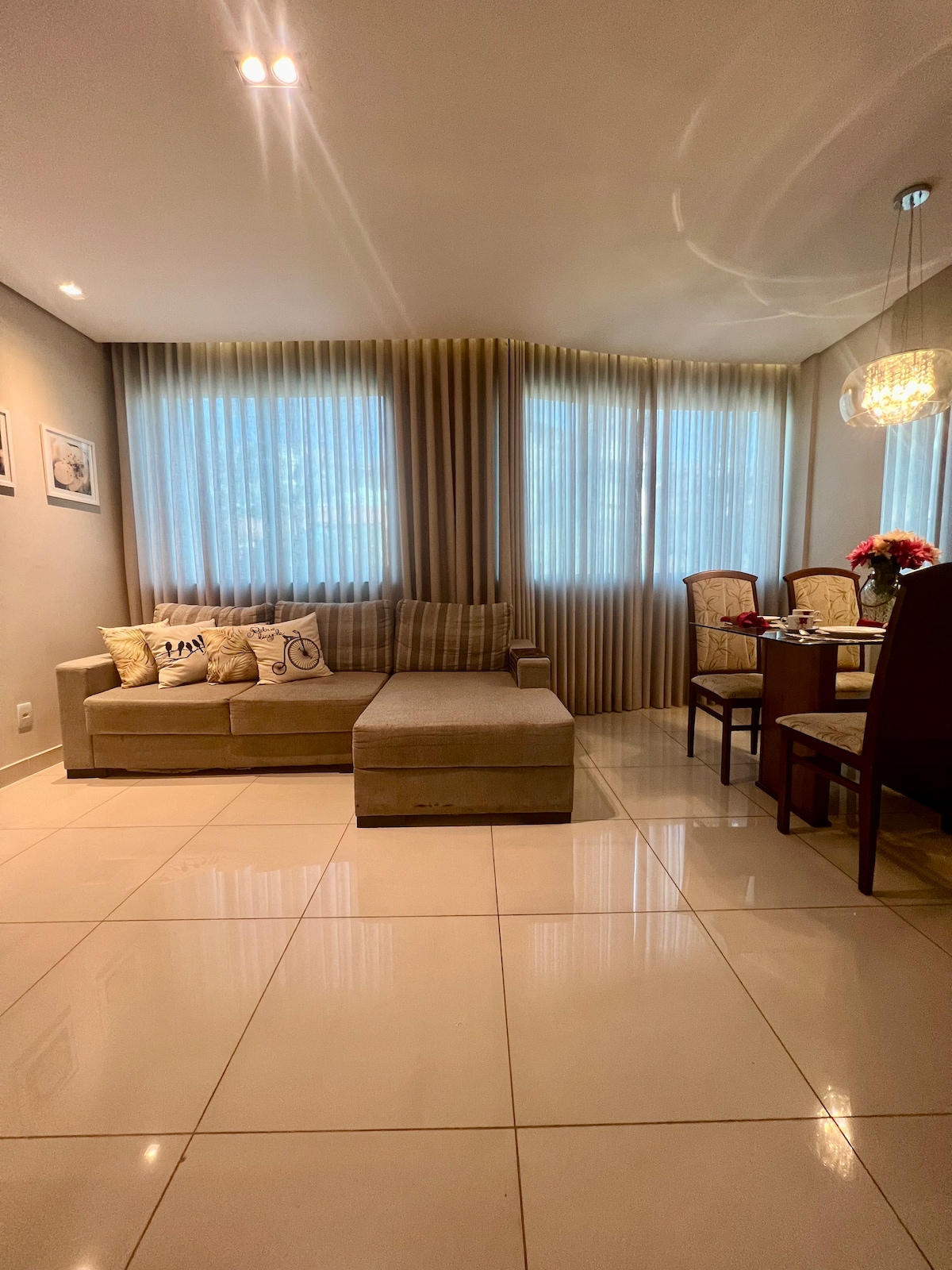 A spacious living area is presented with a light-colored sectional sofa and a dining table accommodating four chairs. Natural light filters through sheer curtains, illuminating the glossy tiled floor and subtle wall decorations, creating a calm and inviting atmosphere.