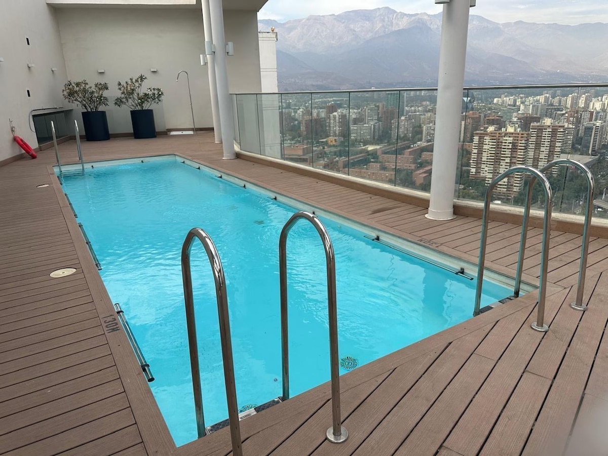 An outdoor pool is surrounded by a wooden deck, offering a clear view of the city skyline and mountains in the background. The pool features stainless steel railings for easy access, and the turquoise water is inviting against the neutral tones of the deck.