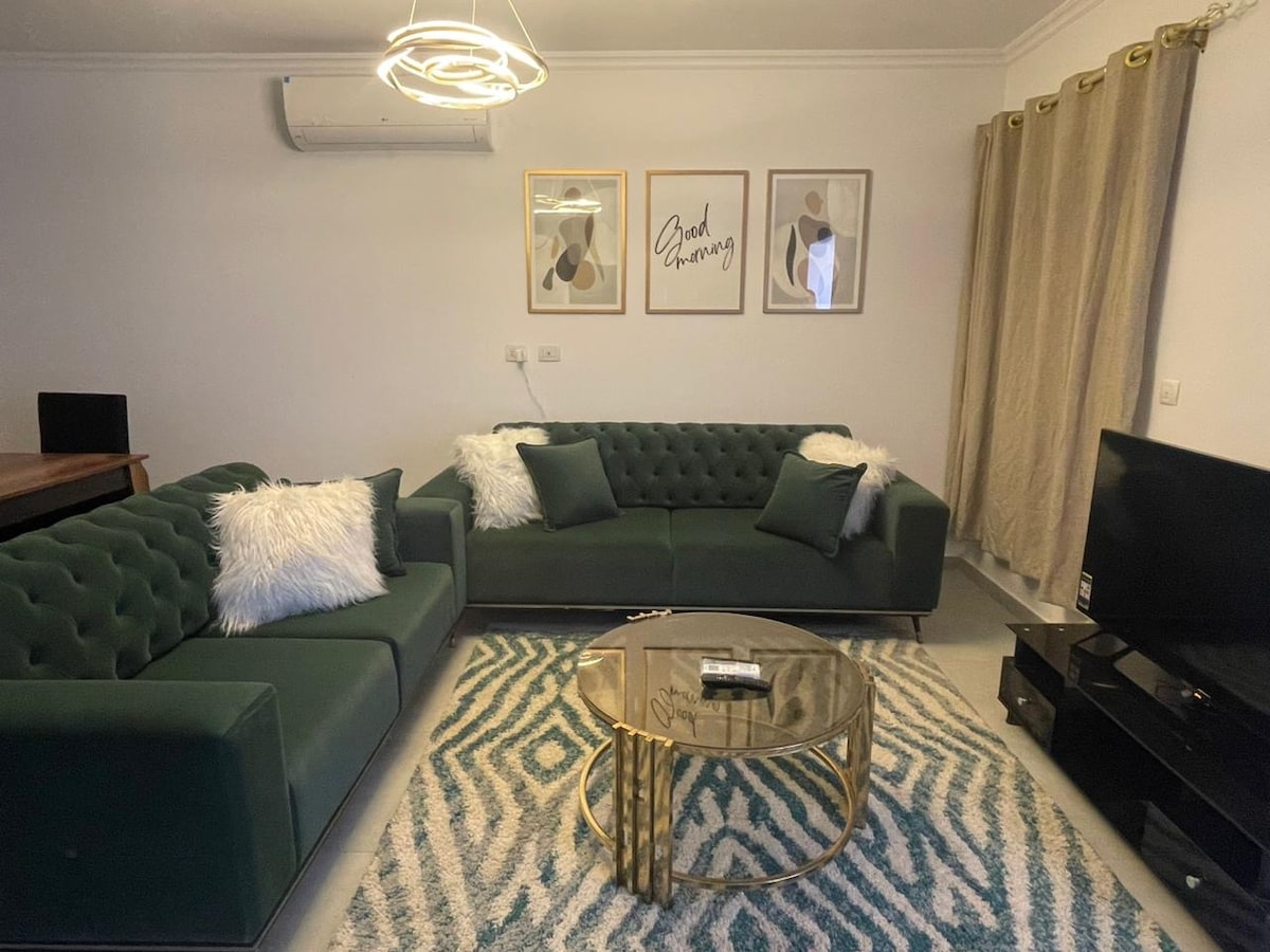 A modern living area is presented, featuring two plush green sofas adorned with fluffy white pillows. A circular glass coffee table sits atop a patterned rug, while framed art pieces are displayed on the wall. Soft lighting is provided by a suspended light fixture.