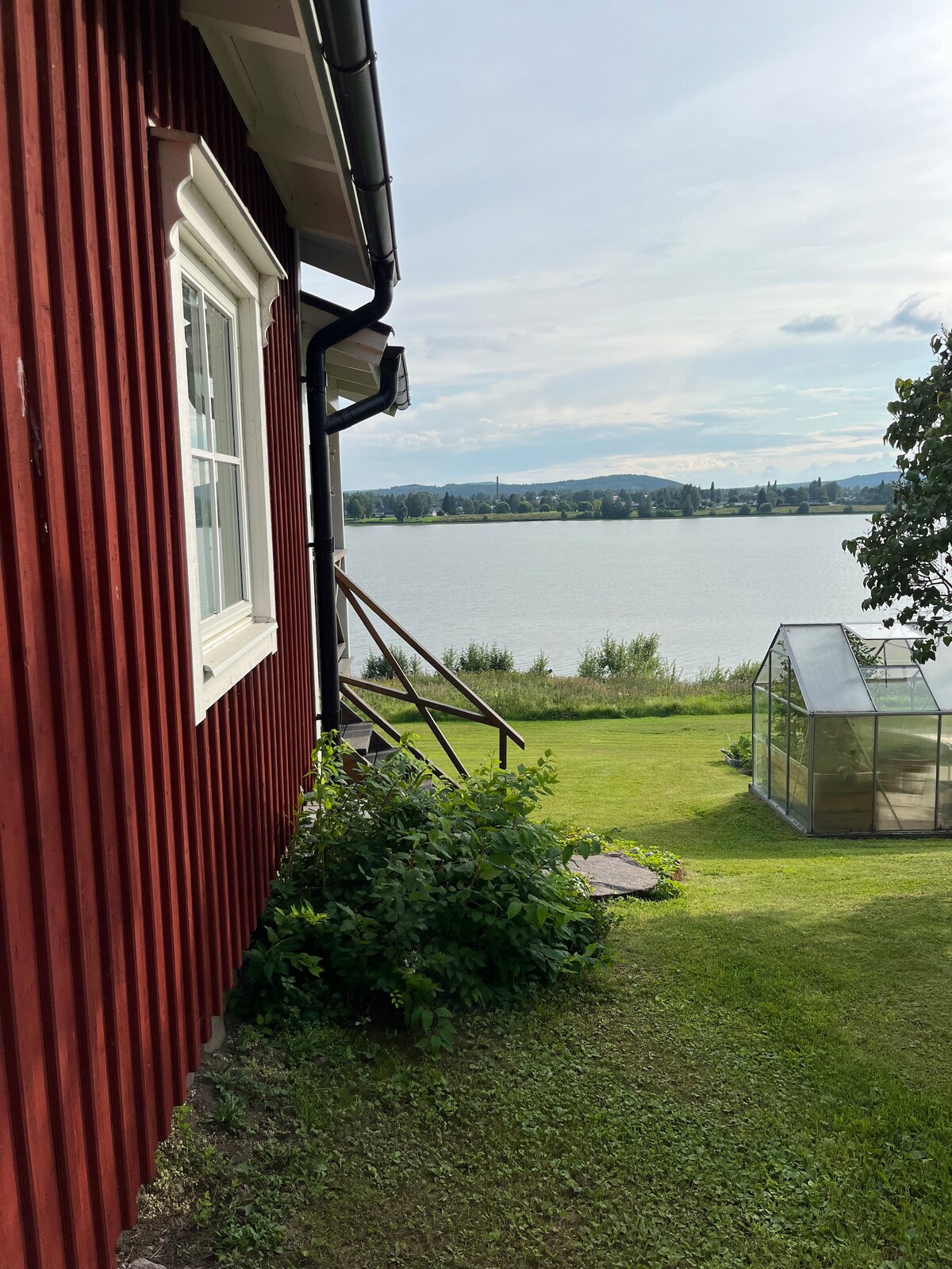 A view from the side of a red wooden cabin is presented, leading out to a green lawn and a serene lake. A greenhouse is visible nearby, set against the backdrop of gentle hills and a cloudy sky.