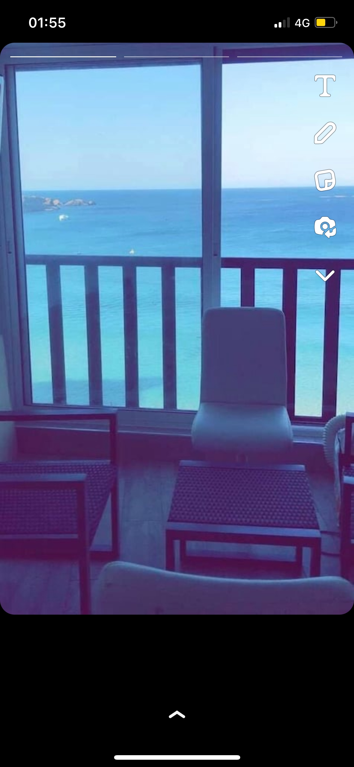A seating area features two chairs and a small table, with large windows offering views of the ocean. The tranquil blue waters can be seen beyond the balcony, while natural light fills the space.