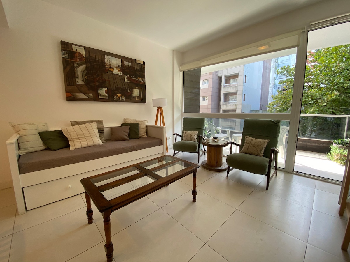A bright living area features a comfortable daybed adorned with neutral cushions, complemented by two green armchairs and a wooden coffee table. Large glass doors allow natural light to fill the space, providing views of the greenery outside.