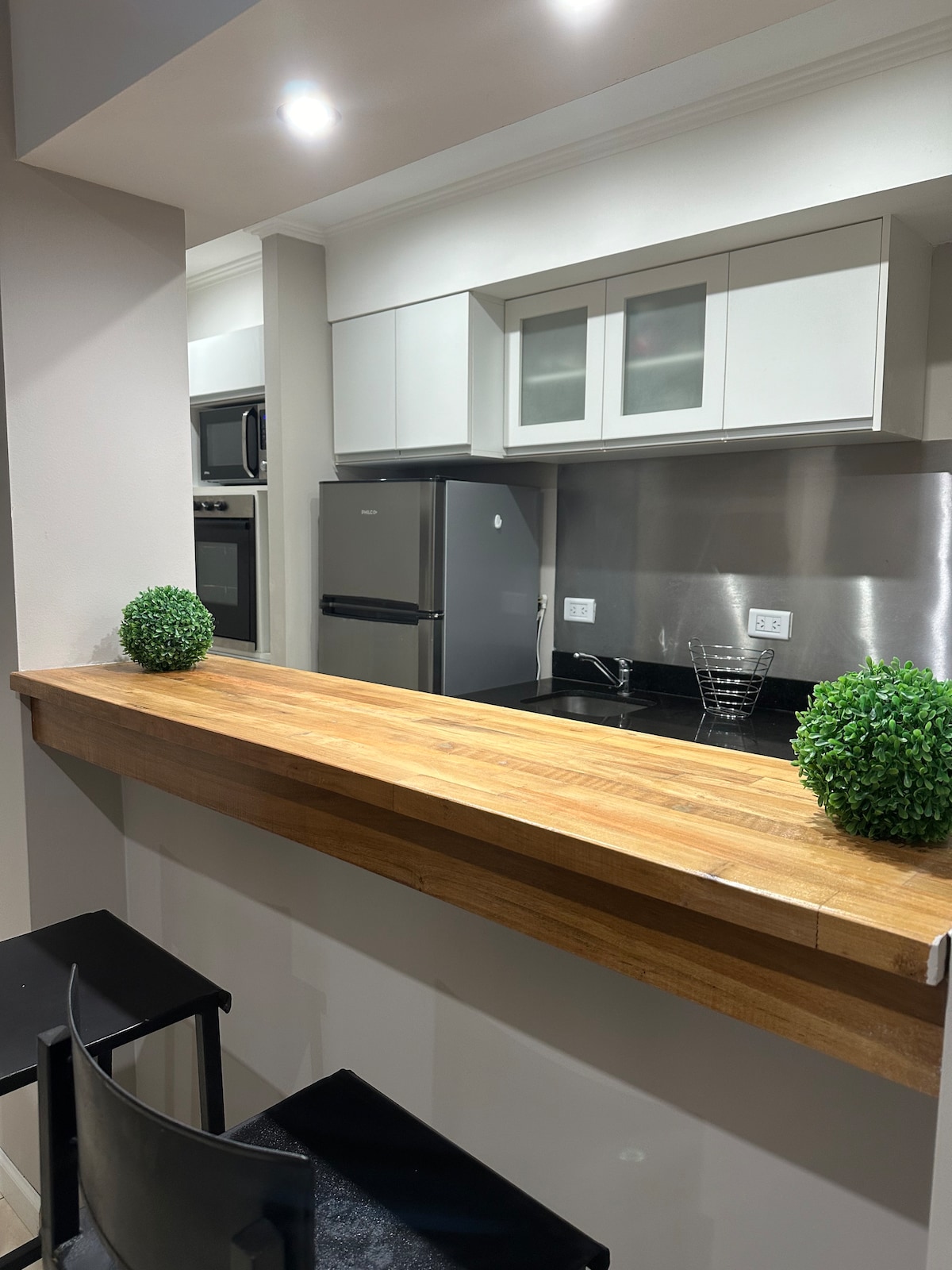 A modern kitchen is visible, featuring sleek cabinetry and stainless steel appliances. A wooden breakfast bar provides seating for two, accented by small green plants on either end. The countertop reflects light, creating a spacious and clean atmosphere.