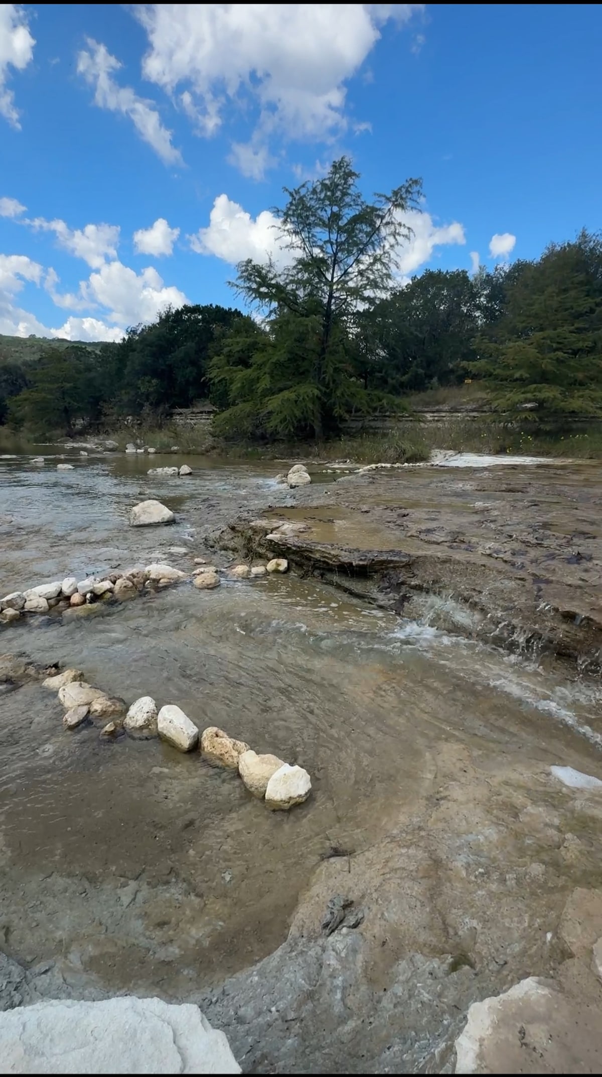 A serene river scene is depicted, featuring gently flowing water over smooth stones. Round stones are arranged along the riverbank, while lush trees provide a backdrop against a bright blue sky adorned with fluffy clouds. The natural setting invites exploration and relaxation.