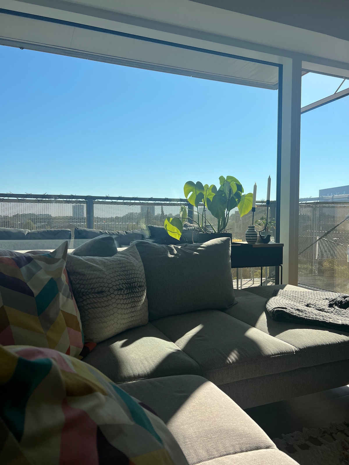 A corner of the living area features a light-filled window showcasing clear blue skies. A sofa adorned with colorful cushions is positioned nearby, alongside a variety of indoor plants that add a touch of greenery to the space.