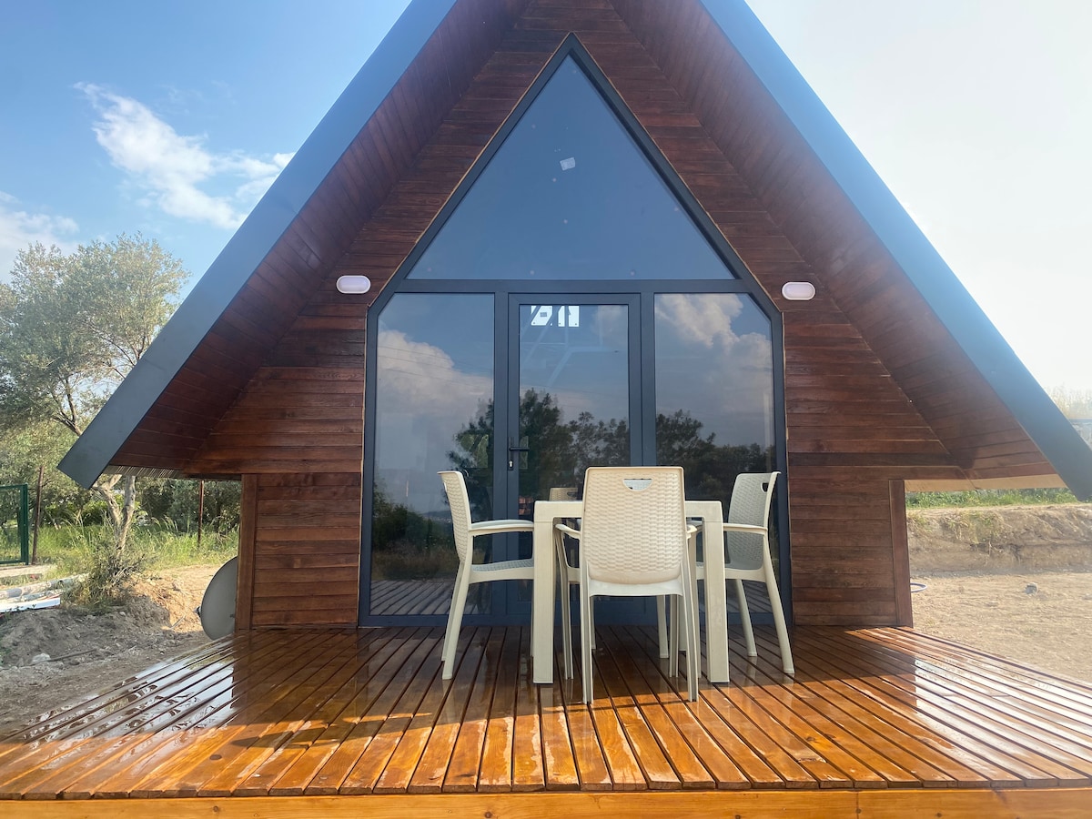 A charming wooden cabin features an A-frame design and large glass doors, allowing natural light to fill the interior. A well-furnished outdoor deck is equipped with a table and four chairs, providing an inviting space for relaxation amidst the surrounding nature.