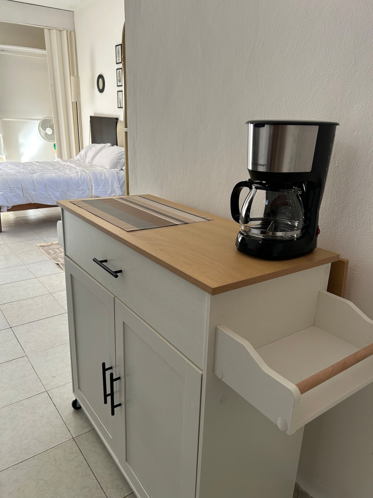 A coffee maker sits on a wooden countertop that is attached to a storage cabinet. The cabinet features a drawer and doors that provide convenient access to supplies. A glimpse of the sleeping area can be seen in the background.