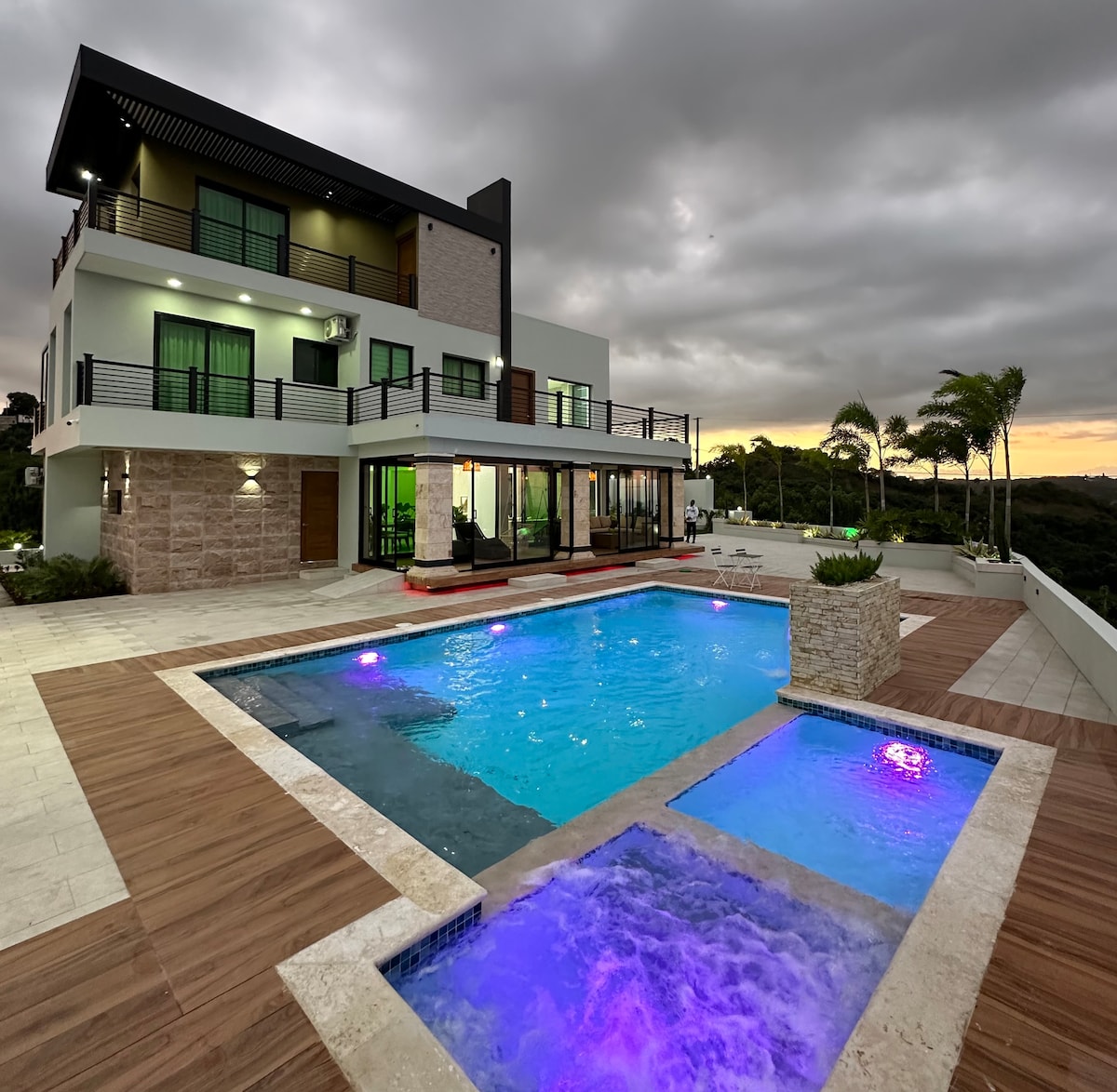 A modern villa is showcased with a spacious outdoor area, featuring a well-lit swimming pool and a jacuzzi. Surrounding the pool is a wooden deck, while the villa's large windows reflect the evening sky, enhancing the view of the lush environment.