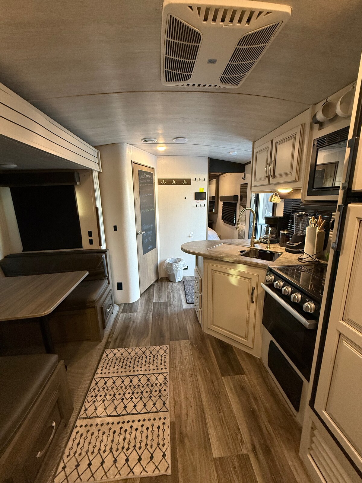 The interior of a travel trailer features a streamlined layout with a cozy dining area and kitchen. Light wood cabinetry contrasts with darker countertops. A distinctive rug adds texture to the floor, while natural light enters through strategically placed windows.