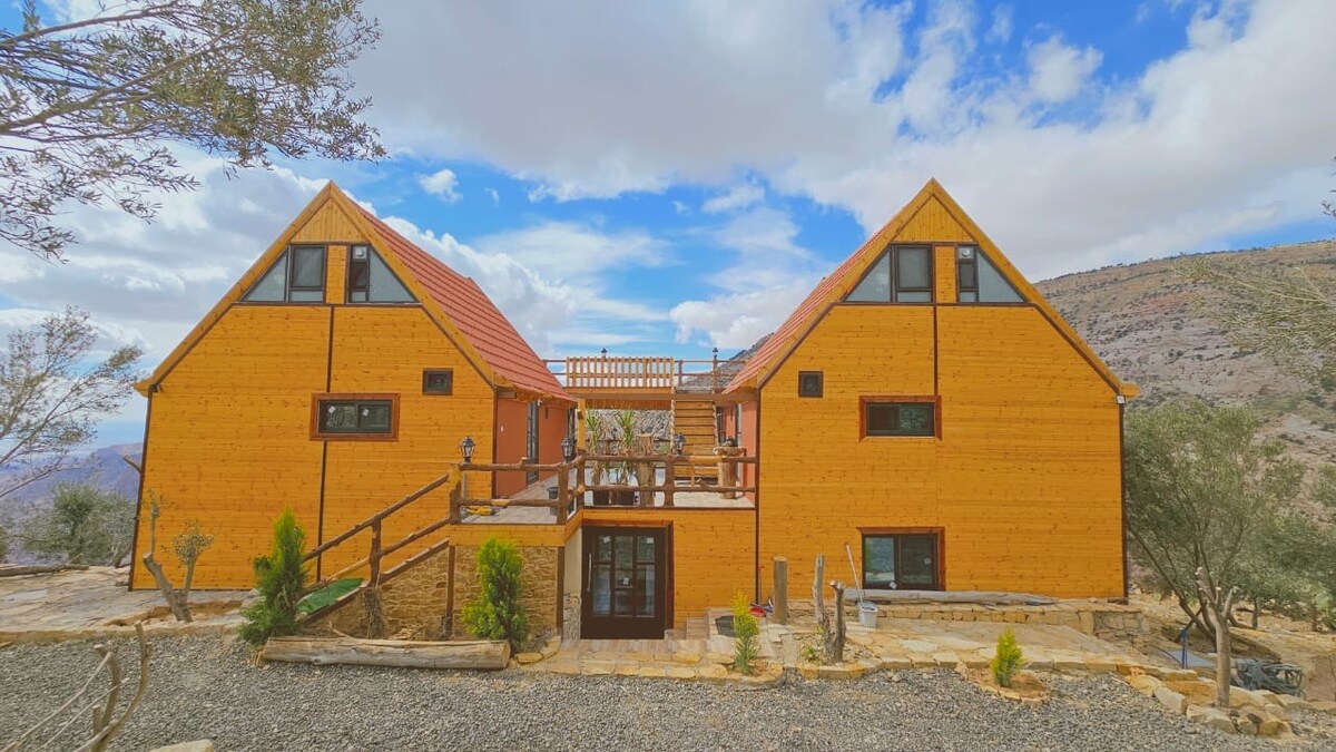 A two-story building with a striking orange exterior features a symmetrical design. A spacious deck is accessible from the upper level, surrounded by natural landscaping. The structure is framed by distant mountains under a partly cloudy sky, enhancing its serene setting.