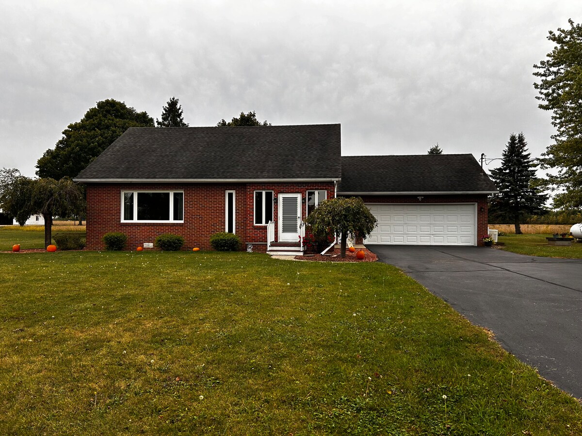 A charming single-story brick home is shown, with a well-maintained lawn featuring scattered pumpkins. A driveway leads to a two-car garage, and a welcoming front entry with steps is visible. The property is surrounded by trees and open fields under an overcast sky.