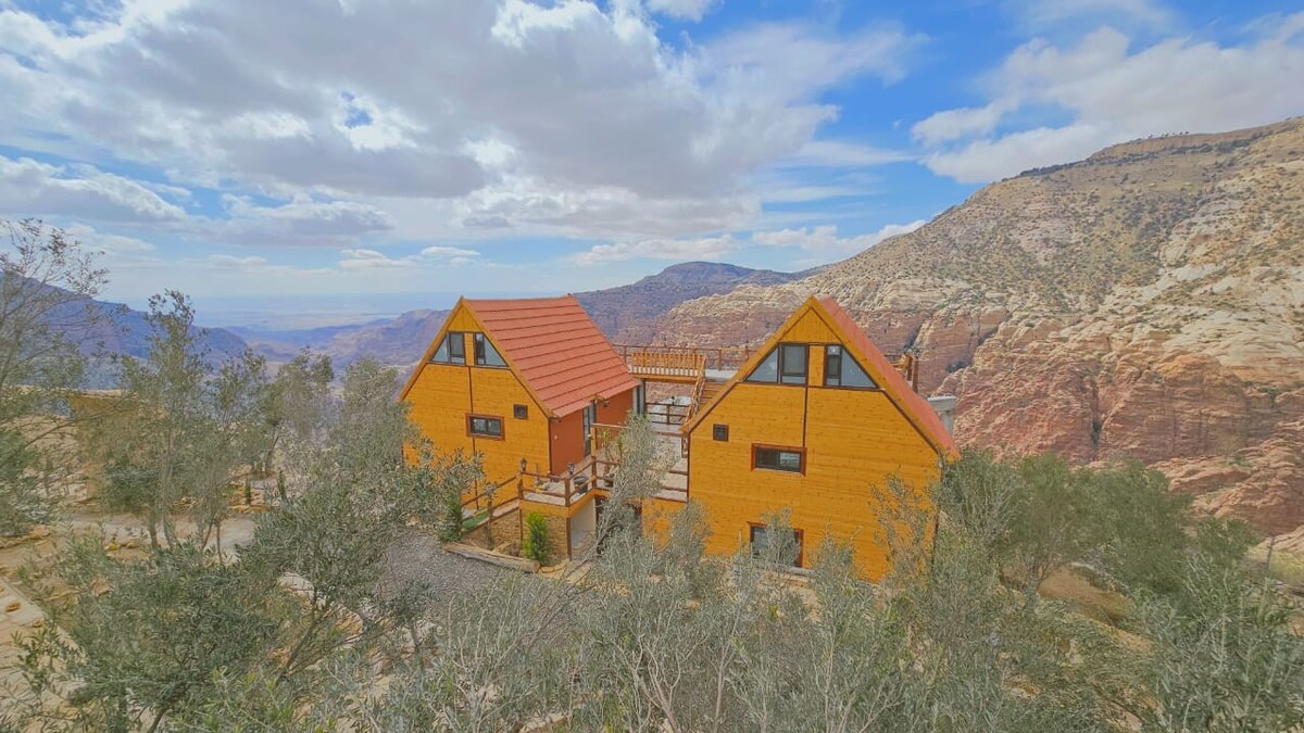 Two charming wooden cabins are positioned side by side, featuring gabled roofs and expansive balconies. Surrounding vegetation includes scattered shrubs and trees. The mountainous landscape in the background highlights a range of earthy tones under a partly cloudy sky.