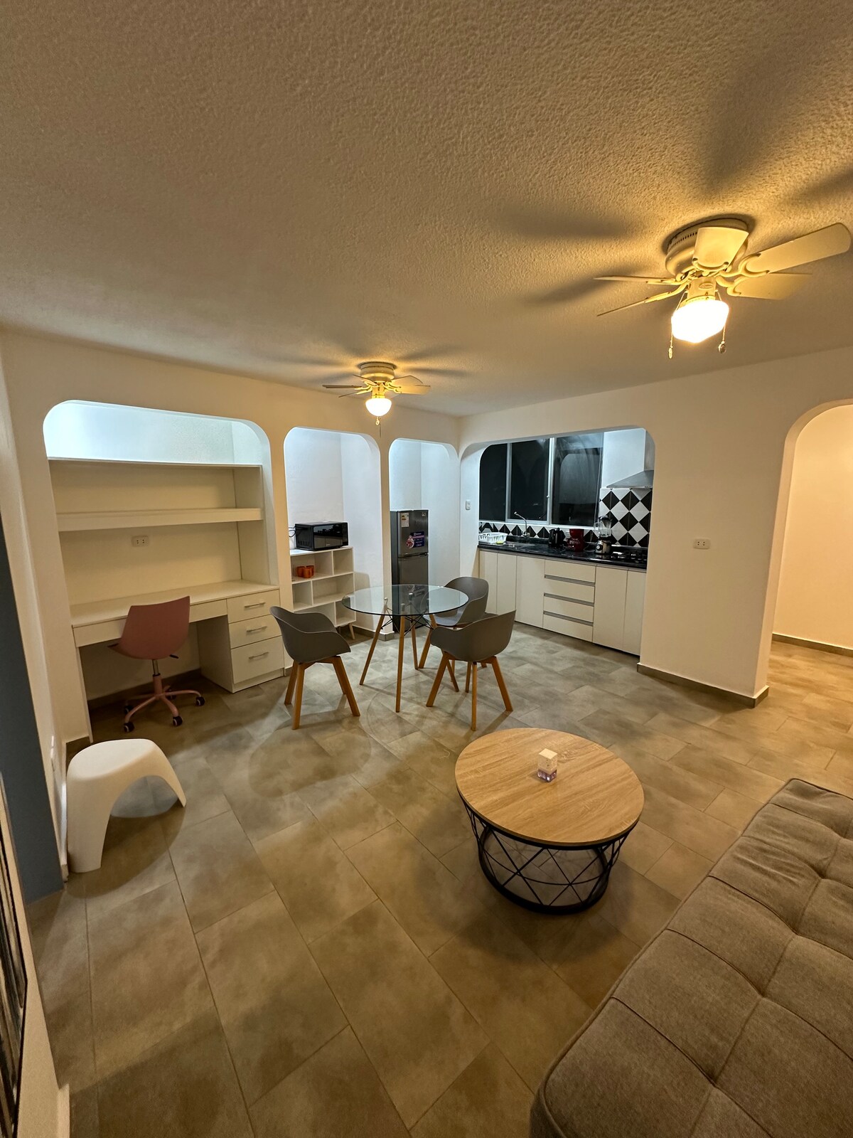 A modern living space is shown featuring a round table surrounded by four chairs. A cozy couch is positioned nearby, with a coffee table in the center. The area includes a workstation with a chair and is illuminated by ceiling fans, enhancing the open, airy feel.