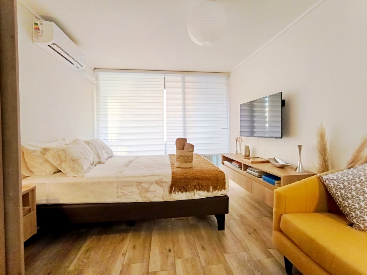A spacious sleeping area features a comfortable bed adorned with neutral-toned linens. Soft light filters through partially closed blinds. A modern television is mounted on the wall, accompanied by a wooden media console. A bright yellow armchair adds a pop of color to the room.