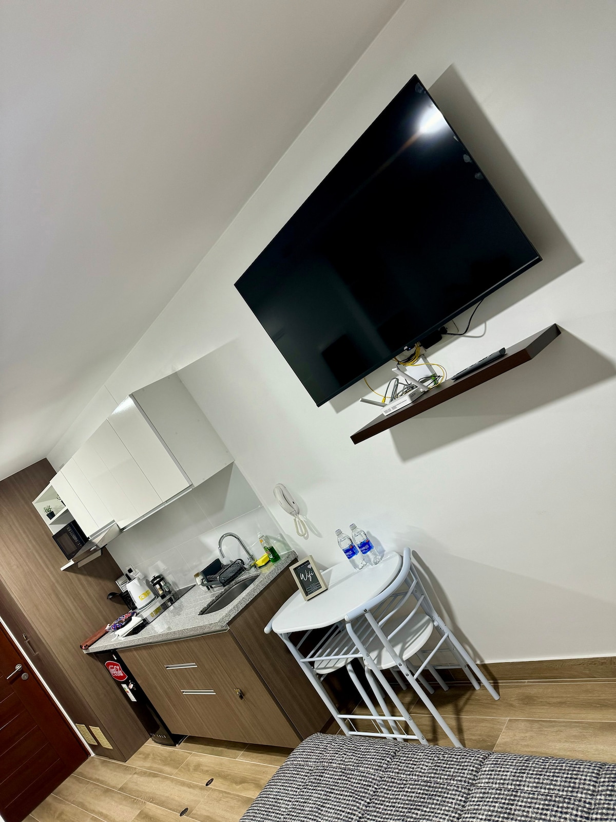 The image displays a modern kitchenette equipped with essential appliances, including a stove and sink. Above, a wall-mounted television is visible, alongside a minimalistic dining area with two chairs and a small table. Neutral tones and wooden flooring contribute to a contemporary aesthetic.