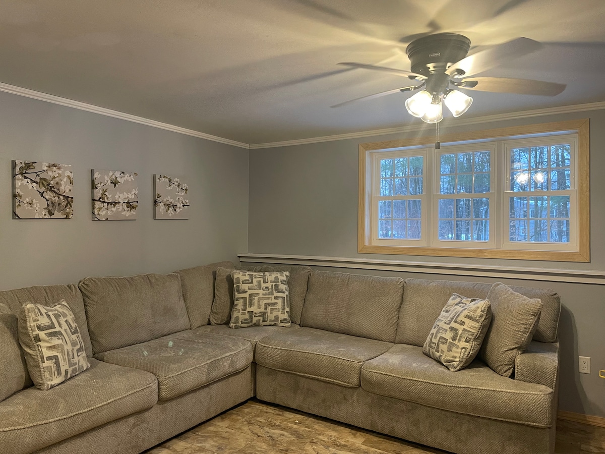 A spacious corner sectional sofa is featured in a softly lit living area. Three pieces of wall art with floral designs complement the light grey walls, while a large window allows natural light to fill the space, framed by white trim.