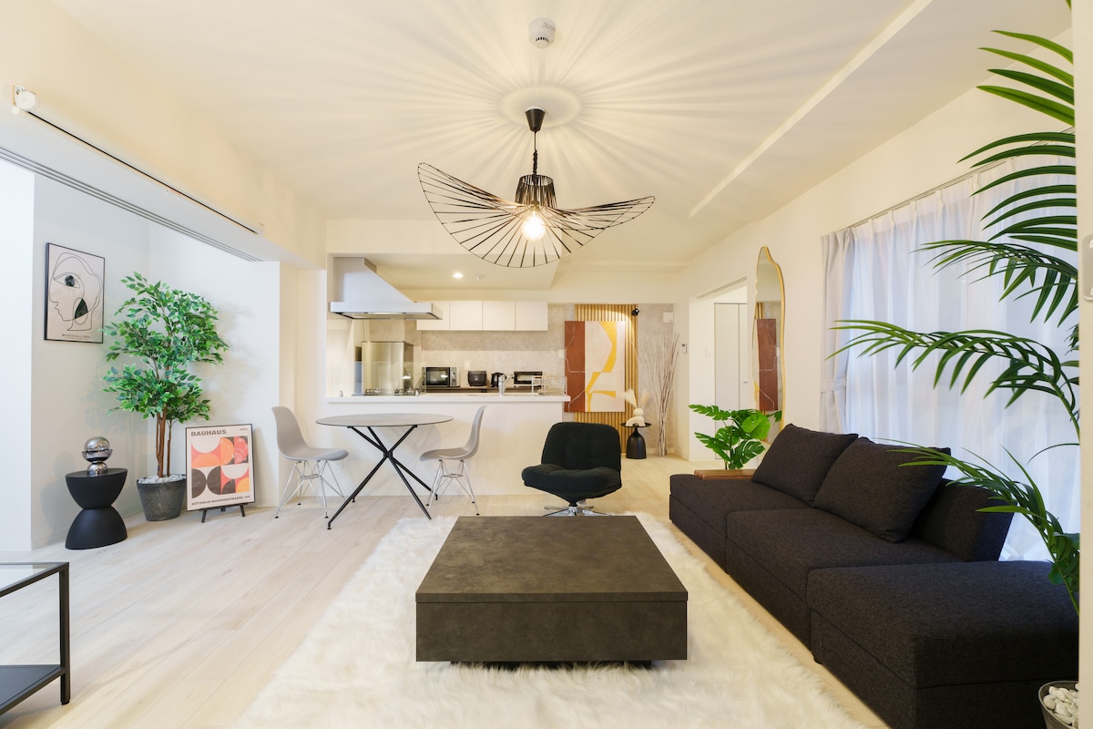 A spacious living area features a modern design with a dark sectional sofa and a stylish coffee table on a soft rug. Large windows allow natural light to brighten the room, complemented by indoor plants and a decorative lamp hanging from the ceiling.