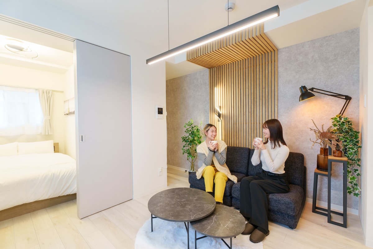 A modern living area features a cozy couch against a decorative wooden wall. Two guests enjoy beverages, surrounded by potted plants and warm lighting from a stylish lamp. A spacious bed is visible in the background, complemented by soft natural light from a window.