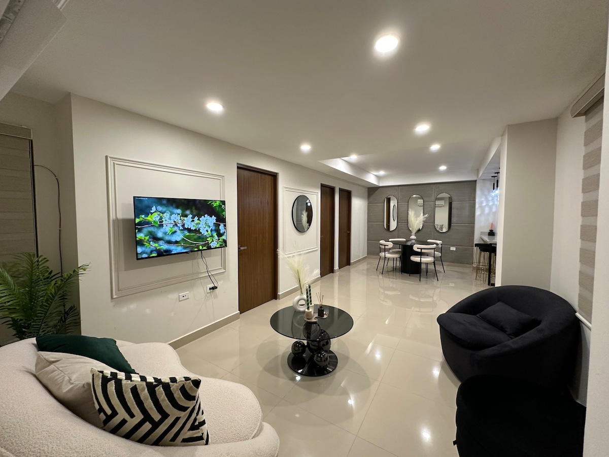 A modern living area is displayed, featuring a comfortable white sofa with green and black accents. A round black table is set at the center, surrounded by stylish dining chairs. Wall-mounted mirrors and artwork contribute to a spacious appearance, while soft lighting enhances the ambiance.