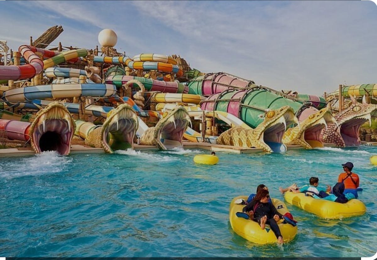 A vibrant water park scene is depicted with colorful water slides cascading into a bright blue pool. Several guests are enjoying themselves in yellow inflatable rings, preparing for a fun ride down the slides that feature playful designs resembling sea creatures.