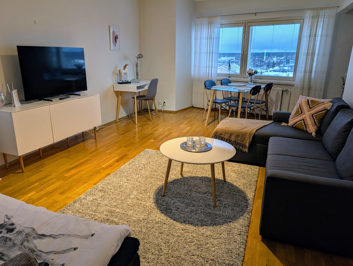 A spacious living area features a large flat-screen television and a cozy sofa, complemented by a light-colored coffee table. A dining table set for four is visible near the window, showcasing city views. Natural light enhances the warm wooden flooring.